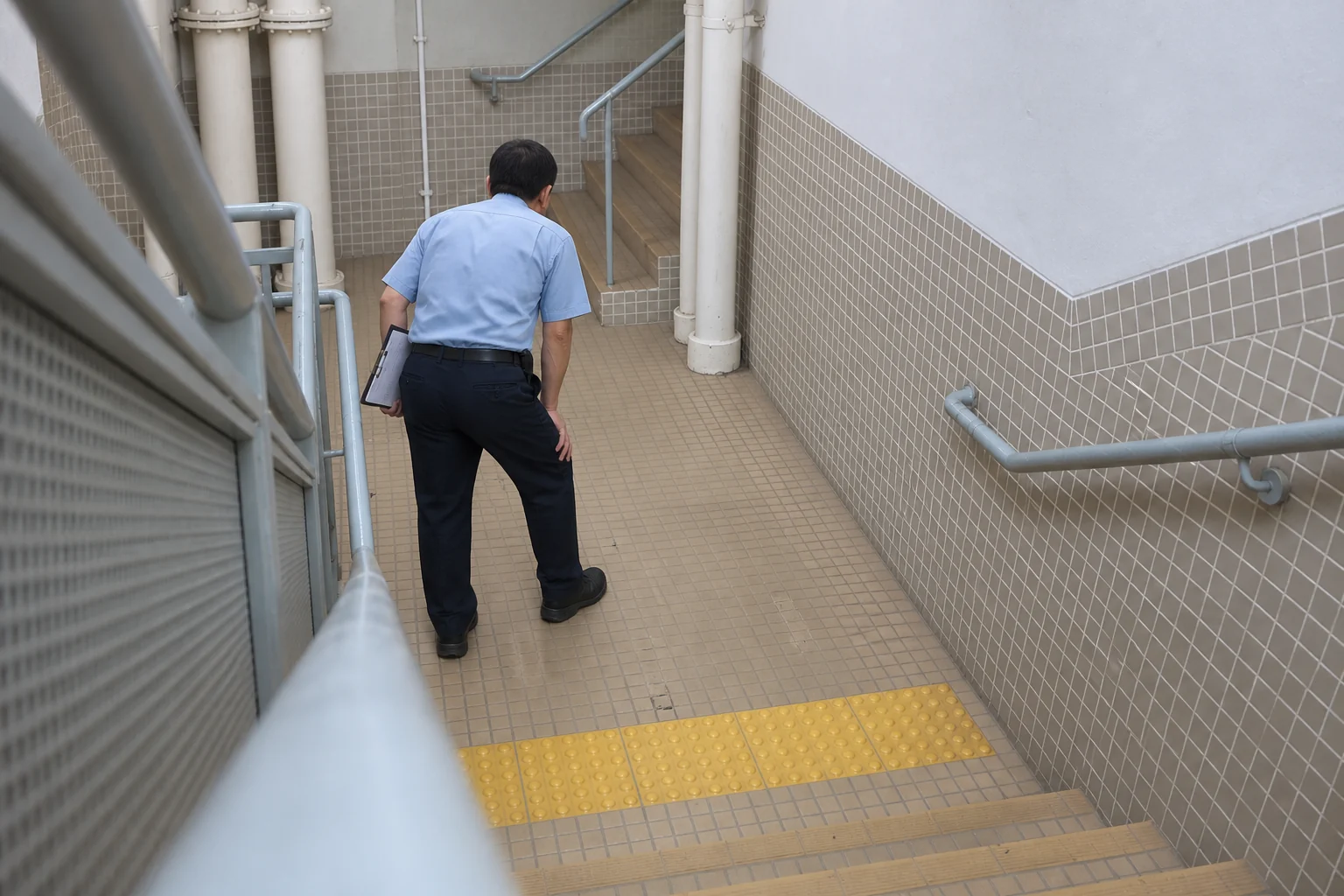階段室で曲げた膝に手をつく警備員の後ろ姿(顔は映っていません)。Stairio の現場画像をもとにした AI イラスト。